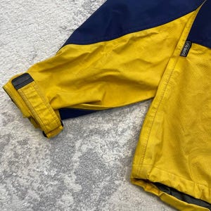 May include: A jacket featuring a color-blocked design in navy blue and bright yellow. The sleeve includes an adjustable cuff with a black strap and buckle. A small black tag with the white text Gore-Tex is stitched onto the yellow fabric.