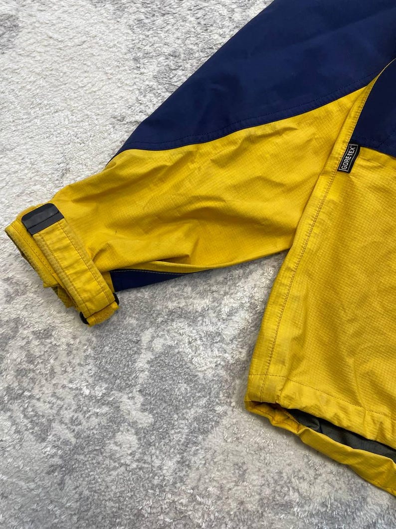 May include: A jacket featuring a color-blocked design in navy blue and bright yellow. The sleeve includes an adjustable cuff with a black strap and buckle. A small black tag with the white text Gore-Tex is stitched onto the yellow fabric.