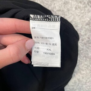 May include: A close-up of a black garment with a white tag attached. The tag displays Chinese characters, fabric composition (95% cotton, 5% spandex), size XXL, and a style number. The garment is held by a person.