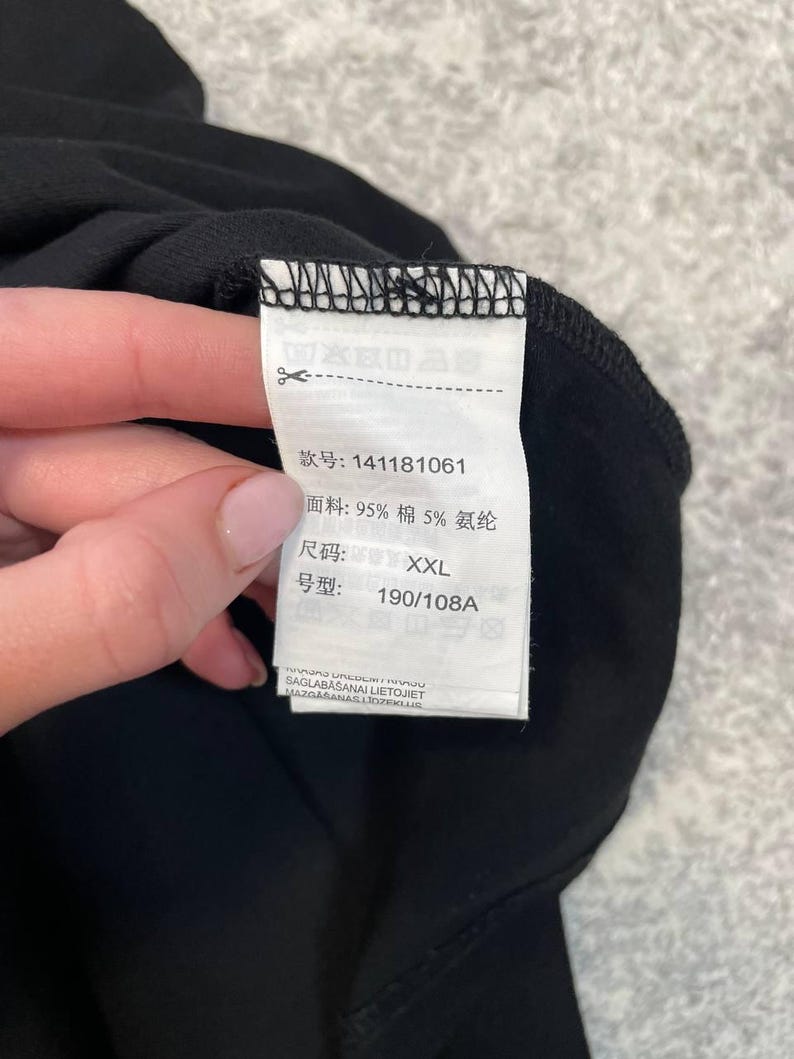 May include: A close-up of a black garment with a white tag attached. The tag displays Chinese characters, fabric composition (95% cotton, 5% spandex), size XXL, and a style number. The garment is held by a person.