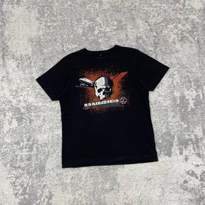 May include: Black t-shirt featuring a graphic design with a skull, a plane, and the word "RAMMSTEIN" in white and orange. The design also includes a red X and a logo. The shirt is made of a soft material.