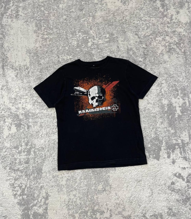 May include: Black t-shirt featuring a graphic design with a skull, a plane, and the word "RAMMSTEIN" in white and orange. The design also includes a red X and a logo. The shirt is made of a soft material.