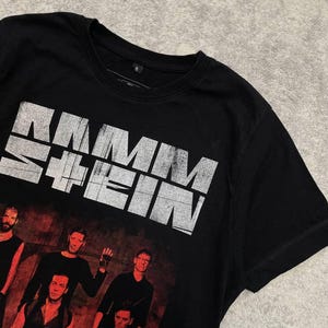 K&ouml;nnte beinhalten: Ein schwarzes Kurzarm-T-Shirt mit dem wei&szlig;en, abgenutzten Schriftzug RAMMSTEIN auf der Brust. Unter dem Text befindet sich ein Grafikdruck von f&uuml;nf Personen in dunkler Kleidung vor einem dunkelroten Hintergrund. Das T-Shirt hat einen Rundhalsausschnitt.