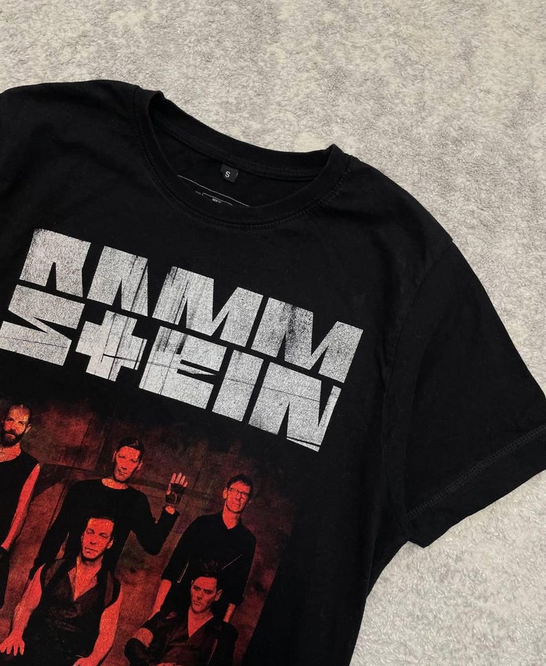 K&ouml;nnte beinhalten: Ein schwarzes Kurzarm-T-Shirt mit dem wei&szlig;en, abgenutzten Schriftzug RAMMSTEIN auf der Brust. Unter dem Text befindet sich ein Grafikdruck von f&uuml;nf Personen in dunkler Kleidung vor einem dunkelroten Hintergrund. Das T-Shirt hat einen Rundhalsausschnitt.