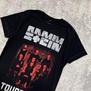 K&ouml;nnte beinhalten: Ein schwarzes Kurzarm-T-Shirt mit einem verwaschenen wei&szlig;en Rammstein-Logo im oberen Bereich. Darunter befindet sich ein rot get&ouml;nter Fotodruck von f&uuml;nf Bandmitgliedern. Der Text TOUR 2017 ist in wei&szlig; am unteren Rand des Designs aufgedruckt.