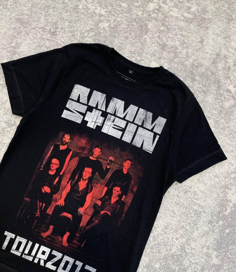 K&ouml;nnte beinhalten: Ein schwarzes Kurzarm-T-Shirt mit einem verwaschenen wei&szlig;en Rammstein-Logo im oberen Bereich. Darunter befindet sich ein rot get&ouml;nter Fotodruck von f&uuml;nf Bandmitgliedern. Der Text TOUR 2017 ist in wei&szlig; am unteren Rand des Designs aufgedruckt.