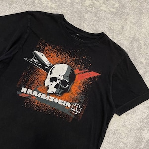 May include: Black t-shirt featuring a skull graphic with orange and white accents. The design includes a stylized airplane and the band name "Rammstein" in white lettering. The shirt has a crew neck.