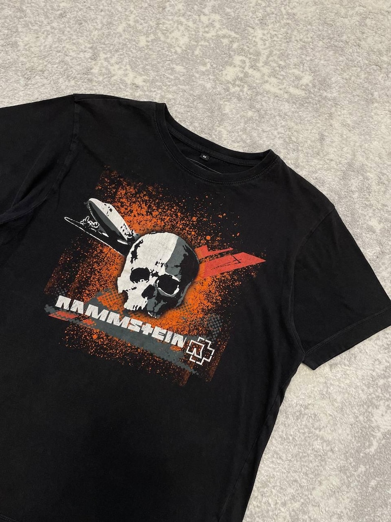 May include: Black t-shirt featuring a skull graphic with orange and white accents. The design includes a stylized airplane and the band name "Rammstein" in white lettering. The shirt has a crew neck.