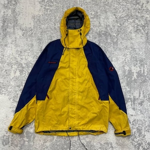May include: A yellow and navy blue hooded jacket featuring a front zipper closure, adjustable drawstrings at the hood and hem, and adjustable velcro cuffs. The Mammut brand logo is visible in red on the chest and on the upper left sleeve.