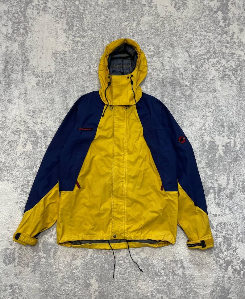 May include: A yellow and navy blue hooded jacket featuring a front zipper closure, adjustable drawstrings at the hood and hem, and adjustable velcro cuffs. The Mammut brand logo is visible in red on the chest and on the upper left sleeve.