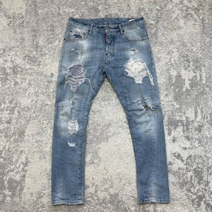 May include: Light blue denim jeans featuring a distressed finish with multiple rips and abrasions. The front includes a blue and white striped fabric patch on the left thigh and a white polka-dot fabric patch on the right thigh, with a button fly.