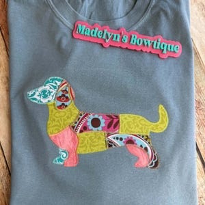May include: A slate blue t-shirt featuring a colorful patchwork-style dachshund applique with various floral and paisley patterns in shades of green, pink, and teal. The text Madelyn's Bowtique is printed in teal on a pink rectangular background.