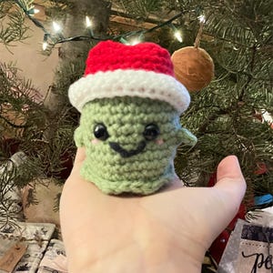 May include: A crocheted green creature wearing a red and white Santa hat. The handmade ornament has black eyes and a smiling mouth. The background includes a Christmas tree with lights and ornaments.