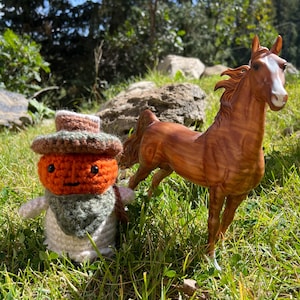 May include: A brown ceramic horse figurine stands in a grassy area next to a crocheted pumpkin doll wearing a hat and scarf. The horse has a white blaze on its face and is posed with its front legs raised. The doll has an orange face and a green scarf.