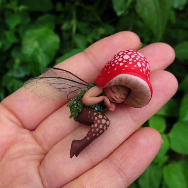 May include: A small figurine of a sleeping creature with a red mushroom cap hat featuring white spots, translucent insect-like wings, a green moss-textured torso, and brown legs with light-colored spots, resting in a palm-sized scale.