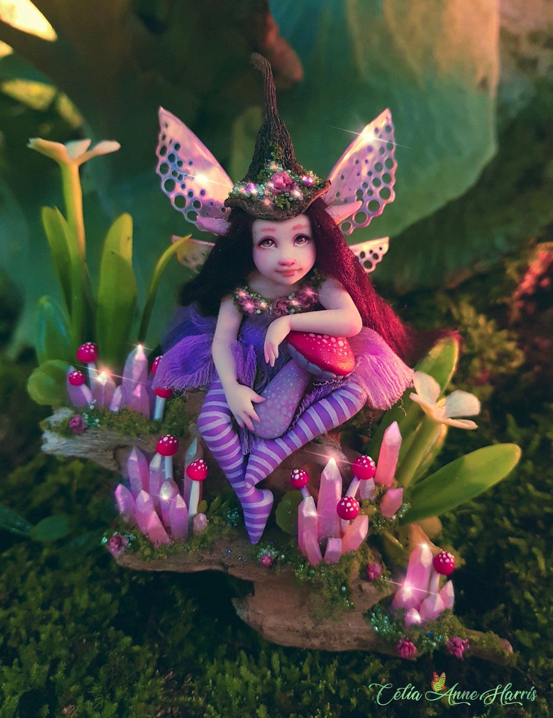 May include: A fairy figurine with dark hair, wearing a pointed hat and purple striped leggings, sitting on a piece of wood adorned with pink crystals and small red mushrooms. The figure has translucent wings and is surrounded by green foliage.