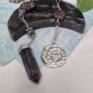 May include: A silver-toned metal chain features a dark purple hexagonal crystal point pendant and a circular charm depicting a sun and crescent moon. The chain is accented with small purple beads and round decorative discs with intricate patterns.