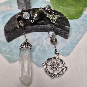 May include: A silver-toned chain pendulum featuring a clear quartz crystal point capped with rhinestones, a dark heart-shaped bead, and a circular compass charm. The piece includes faceted grey beads and intricate metal connectors on a dark chain.