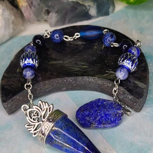 Peut inclure: Un pendule composé d'un pendentif conique en lapis-lazuli bleu profond avec un capuchon en forme de fleur de lotus argentée. La chaîne comporte diverses perles bleues, dont du verre dépoli, de la pierre marbrée et des entretoises argentées.