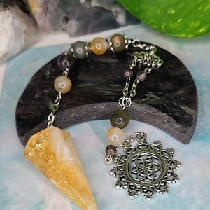 May include: A citrine crystal pendulum with a silver chain and a decorative pendant featuring a flower of life design. The pendulum is paired with a beaded necklace of various earth-toned stones.