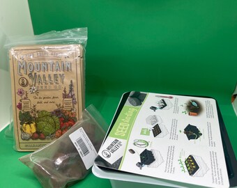 May include: A seed starting kit with a white tray, a black lid with instructions, and a packet of soil. A tin with the words "Mountain Valley Seeds" and a bag of seeds are also present. The kit is on a green surface.