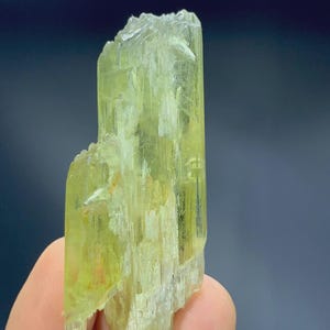 Könnte beinhalten: Ein durchscheinender, hellgelb-grüner Kristall. Die Oberfläche des Minerals ist rau und strukturiert, mit sichtbaren Streifen und einer kristallinen Struktur. Die Form ist länglich und unregelmäßig, mit einem natürlichen, unpolierten Aussehen.