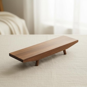 Könnte beinhalten: Ein rechteckiges Serviertablett aus Holz mit warmbraunem Finish. Das Tablett wird von drei kurzen, zylindrischen Beinen getragen. Das Design ist minimalistisch und modern, geeignet zum Präsentieren von Gegenständen oder zum Servieren von Speisen. Das Tablett ist ca. 30 cm lang.