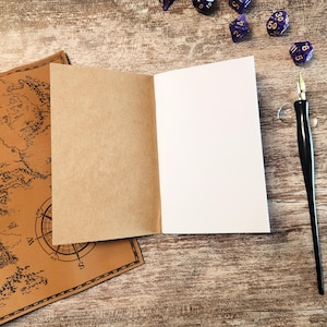 May include: An open notebook with blank white pages and a brown cover sits on a wooden surface. A map with a compass rose is visible on the left. A black pen and several purple dice are also present, suggesting a tabletop gaming theme.