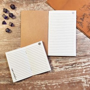 May include: Two open notebooks with tan covers and white lined pages. The smaller notebook features a small 20-sided die icon at the top right and a vertical column of various polyhedral die icons on the left. Seven dark purple dice are nearby.