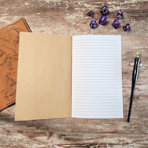 May include: An open, lined notebook with a kraft paper cover sits on a wooden surface, next to a black calligraphy pen and a set of purple dice. A leather-bound journal with a map design is partially visible.
