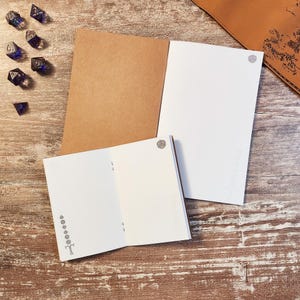 May include: Two open notebooks with brown covers and white pages, one larger than the other. The pages feature small gray icons of polyhedral dice. Seven dark blue translucent polyhedral dice are scattered to the left of the notebooks.
