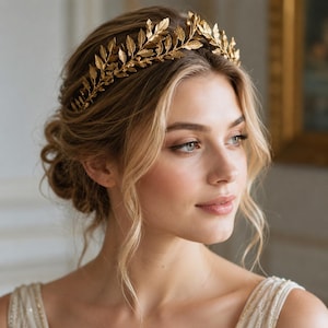 May include: A gold-colored leaf crown, worn on the head. The crown is made of metal and features detailed leaf designs. The woman has blonde hair and is wearing a white dress. The background is a soft, neutral color.