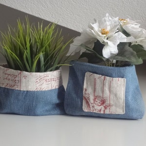 Peut inclure: Deux pots en tissu denim bleu avec des doublures en tissu blanc. Un pot a une doublure en tissu blanc avec des lettres rouges. L'autre pot a une doublure en tissu blanc avec une petite image d'un ange. Les deux pots sont remplis de verdure.