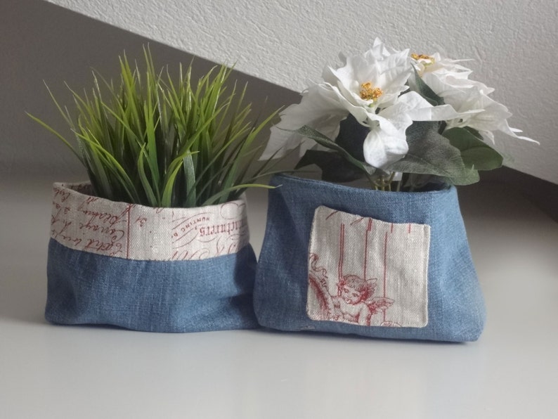 Peut inclure: Deux pots en tissu denim bleu avec des doublures en tissu blanc. Un pot a une doublure en tissu blanc avec des lettres rouges. L'autre pot a une doublure en tissu blanc avec une petite image d'un ange. Les deux pots sont remplis de verdure.