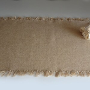 May include: A rectangular burlap placemat with a fringed edge. The placemat is a light brown color. A small bundle of cream-colored fabric roses rests on the right side of the placemat.
