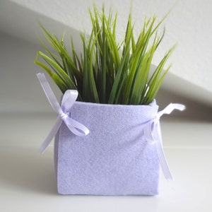May include: A small, lavender felt planter with a green artificial plant inside. The planter has two white ribbons tied around it.