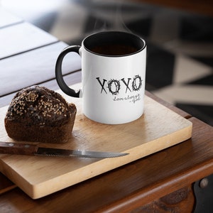 May include: White ceramic coffee mug with a black handle and a black rim. The mug has a black graphic that says "XOXO love always - God" with a crown of thorns design. The mug is sitting on a wooden cutting board with a brown muffin next to it.