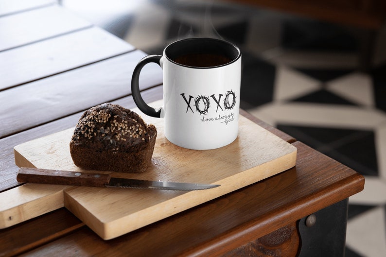 May include: White ceramic coffee mug with a black handle and a black rim. The mug has a black graphic that says "XOXO love always - God" with a crown of thorns design. The mug is sitting on a wooden cutting board with a brown muffin next to it.
