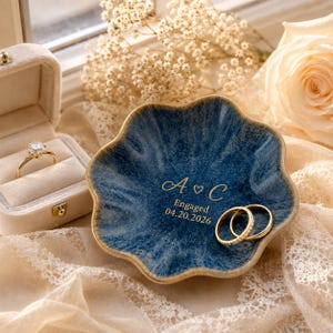 May include: A scalloped, deep blue ceramic ring dish featuring gold-colored text that reads A heart C, Engaged, and 04.20.2026. The dish is accompanied by a gold engagement ring with a solitaire stone and two gold wedding bands.