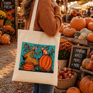 May include: A cream-colored canvas tote bag featuring a colorful illustration of a person in a yellow patterned dress sitting next to a large orange pumpkin with a small pig, surrounded by swirling blue patterns and falling autumn leaves.