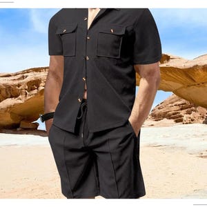 May include: Black short-sleeved shirt and matching shorts. The shirt has two chest pockets and button closures. The shorts have a drawstring waist and vertical seams. The outfit is made of a textured fabric. The background shows a desert landscape.