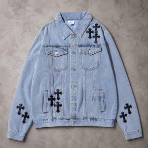 Può includere: Una giacca in denim azzurro con colletto a punta, maniche lunghe e chiusura frontale con bottoni. La giacca è decorata con diverse toppe nere a forma di croce sul petto, sulle maniche e sui pannelli frontali, e include due tasche sul petto.
