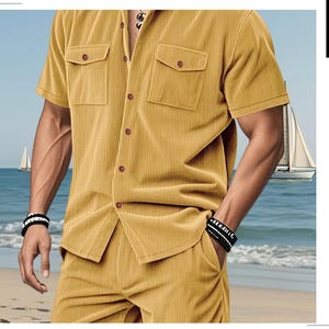 May include: Mustard yellow corduroy shirt and matching shorts set. The short-sleeved shirt has two chest pockets and brown buttons. The shorts have pockets. The background shows a beach with sailboats and the ocean.