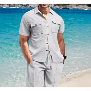 May include: A light gray short-sleeved shirt and shorts set. The shirt has two chest pockets and button closures. The shorts have a drawstring waist. The outfit is made of a textured fabric, perfect for a casual summer look.