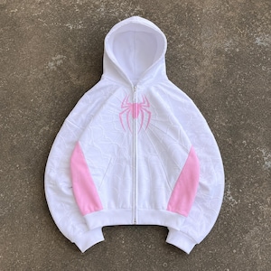 Puede incluir: Una sudadera blanca con cremallera, un emblema de araña rosa en el pecho y un sutil patrón de telaraña en relieve en la tela. La prenda incluye paneles laterales de color rosa sólido, mangas largas con puños acanalados y una capucha a juego.