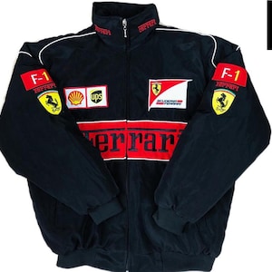 May include: Black zip-up jacket with red and white accents, featuring Ferrari logos and F-1 patches. The jacket has a stand-up collar and ribbed cuffs. The Ferrari logo is prominently displayed across the chest. A stylish piece of outerwear.