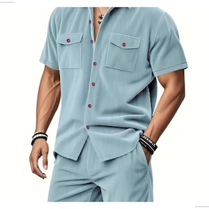 May include: A light blue, short-sleeved shirt and matching shorts set. The shirt has two chest pockets and brown buttons. The fabric has a vertical ribbed texture. The model is wearing black and white bracelets.