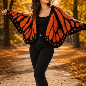 May include: A knitted cardigan featuring a vibrant monarch butterfly wing pattern in bright orange and black. The garment has an open front, wide sleeves, and a textured yarn construction, worn over a black tank top and black denim jeans.
