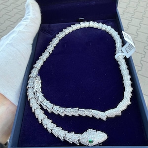 Puede incluir: Un collar con forma de serpiente plateada que presenta un cuerpo segmentado cubierto de pequeñas piedras transparentes. La cabeza de la serpiente tiene una piedra verde como ojo. El collar se presenta en una caja forrada de terciopelo azul.
