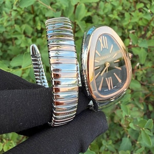 Puede incluir: Un reloj Bvlgari Serpenti con una caja en forma de gota, un bisel de oro rosa y una esfera negra con efecto rayos de sol. El reloj cuenta con una pulsera flexible y enrollable hecha de segmentos metálicos alternos en plata y oro rosa.